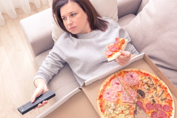 woman eating pizza and holding phone laying on sofa at home