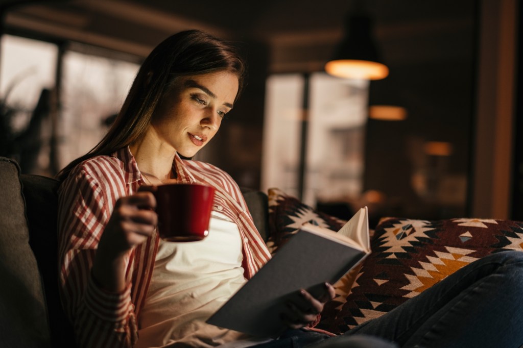 Kobieta w zimowy wieczór czyta książkę siedząc na kanapie z kubkiem w ręce