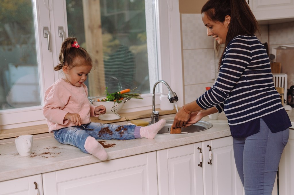 Mała dziewczynka siedzi na kuchennym blacie obok zlewu i robi bałagan, podczas gdy jej matka zmywa naczynia