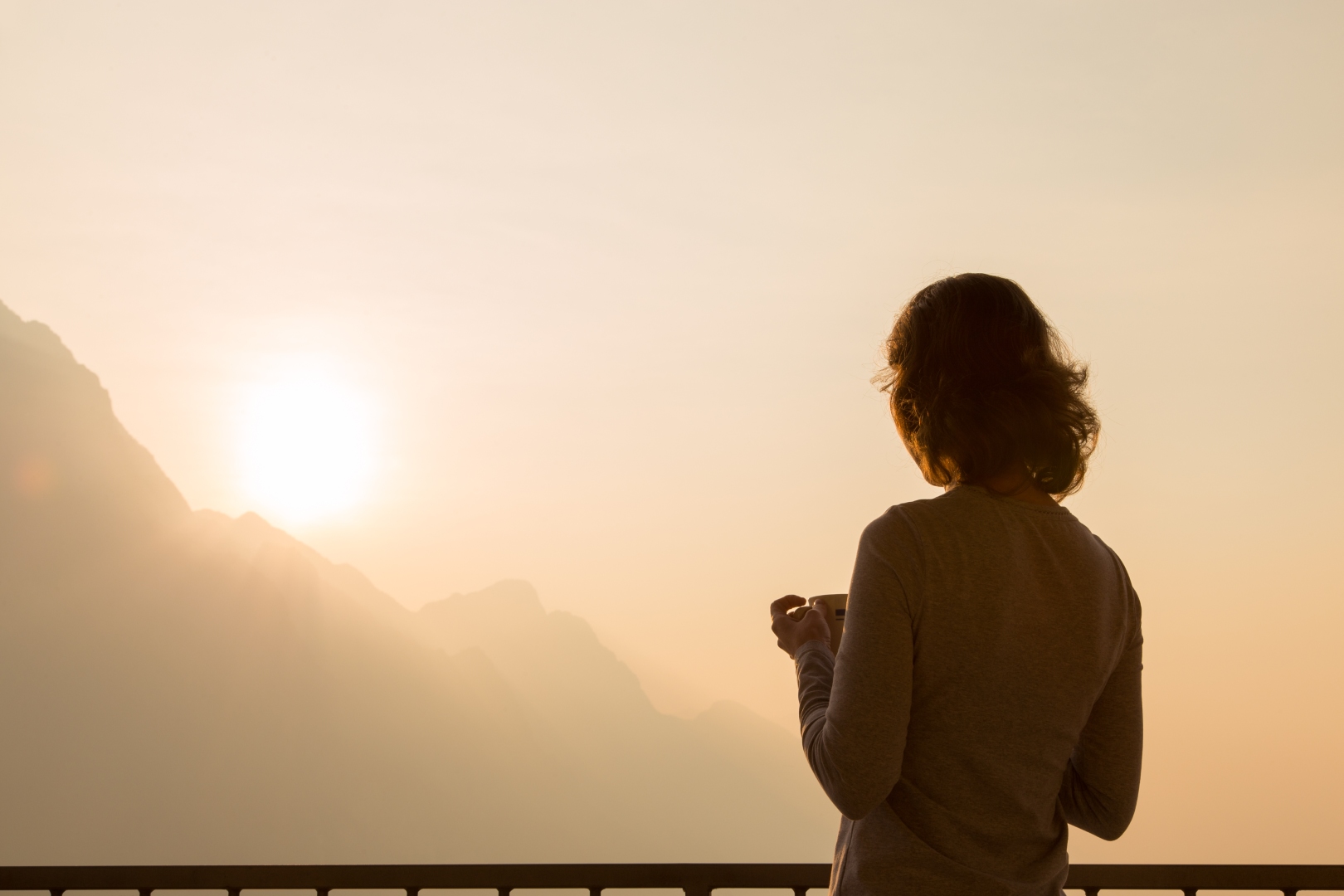 Kobieta z kubkiem kawy wpatruje się w mglisty poranek we wschodzące słońce