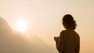 Kobieta z kubkiem kawy wpatruje się w mglisty poranek we wschodzące słońce