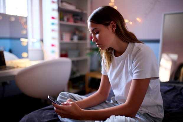 Depressed Teenage Girl Sitting On Bed At Home Looking At Mobile Phone