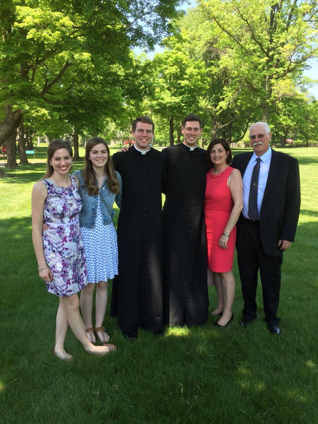 Family posing outside. Two sons in the middle in cassocks.